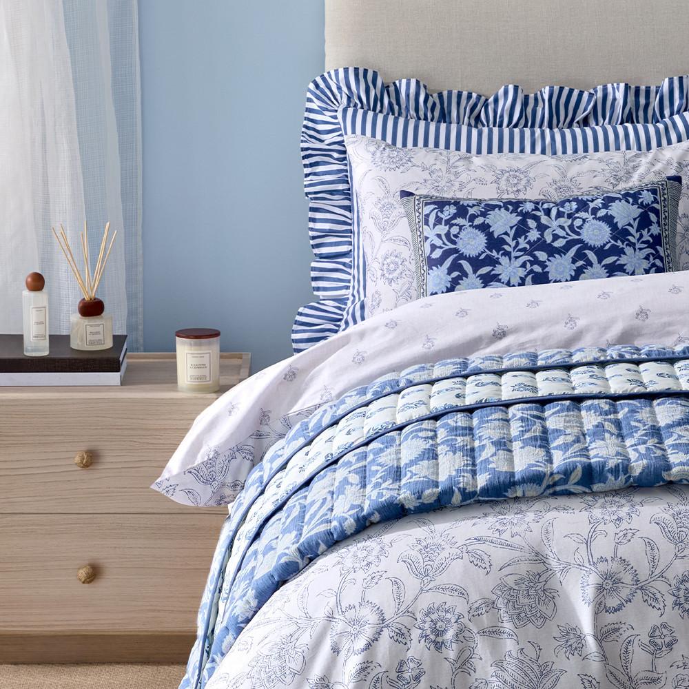 Blau-weiße Bettwäsche mit passenden Kissen mit Blumenmuster und gestreiften Rüschen, daneben ein Diffusor mit Stäbchen und eine Kerze.