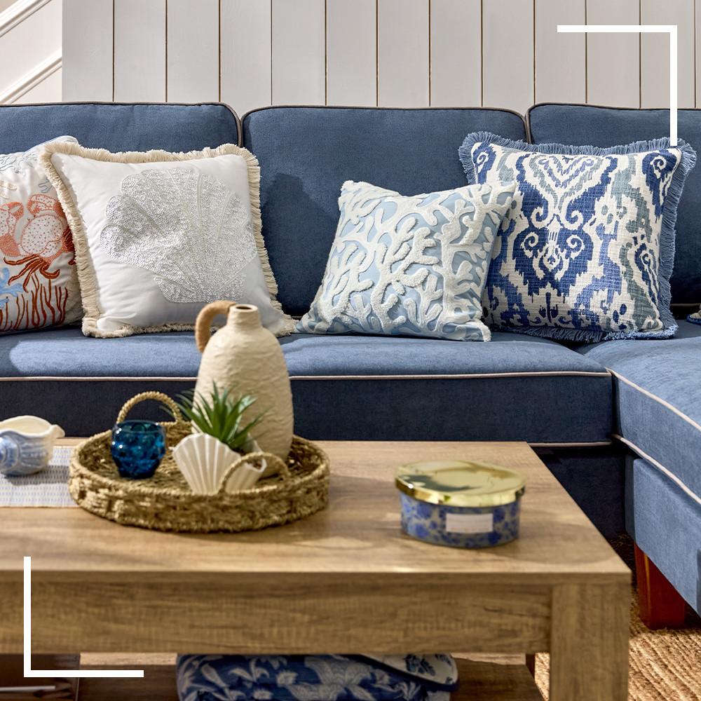 Navy blue sofa with coastal-themed cushions in white and blue patterns, wooden coffee table with a seashell plant pot and vase decor inside a seagrass tray, next to a blue candle with a gold lid.