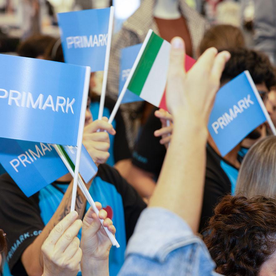People hold blue Primark flags and an Italian flag in a crowd, with hands raised.