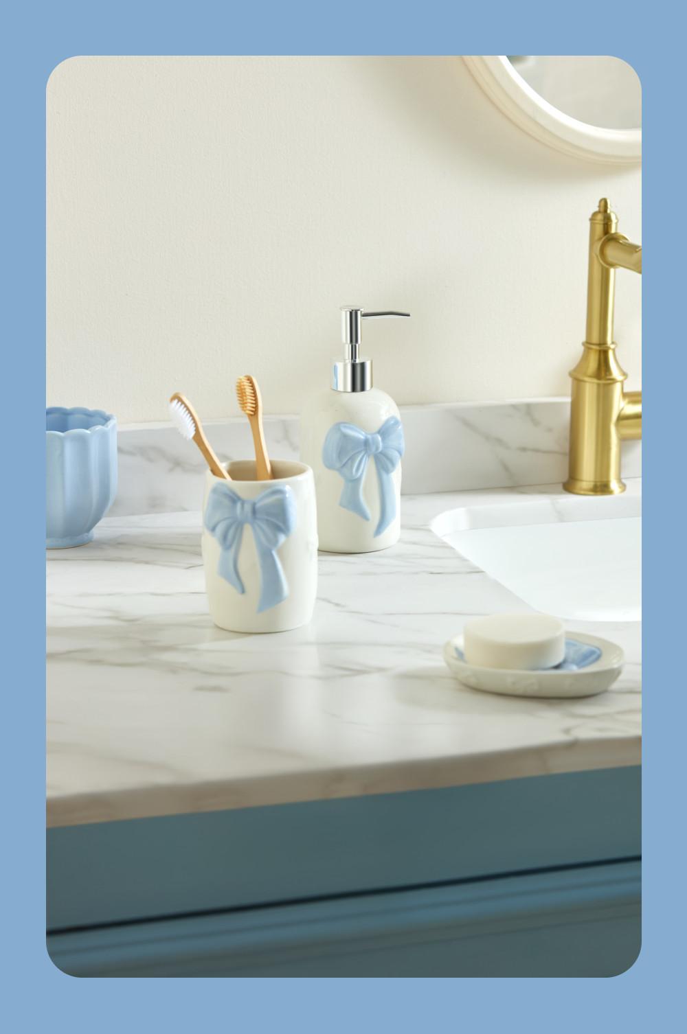 Elegant bathroom set with white ceramic accessories featuring blue bows, including toothbrush holder and soap dispenser on marble counter.