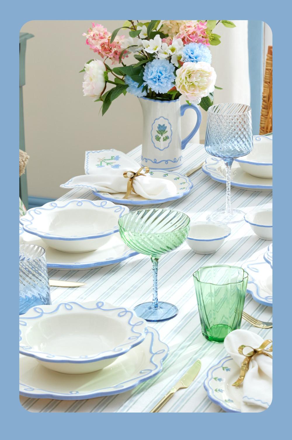 Elegant table setting with blue-rimmed white dinnerware, colourful glassware and floral centrepiece in a jug.