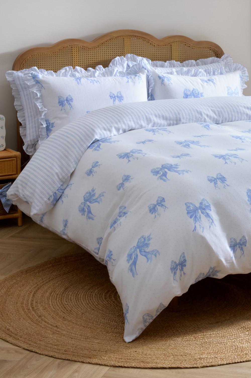 White bedding set with blue bow pattern and ruffled edges on a wooden headboard bed with jute rug.