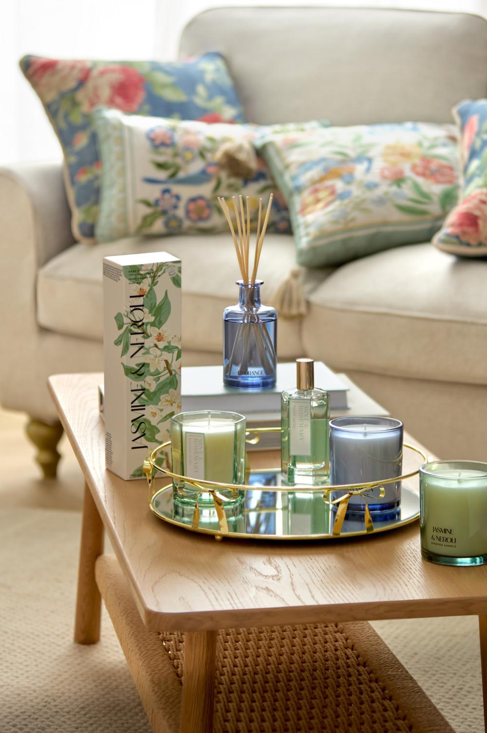 Gold mirrored tray with Jasmine & Neroli fragrance products on wooden coffee table near floral cushions.