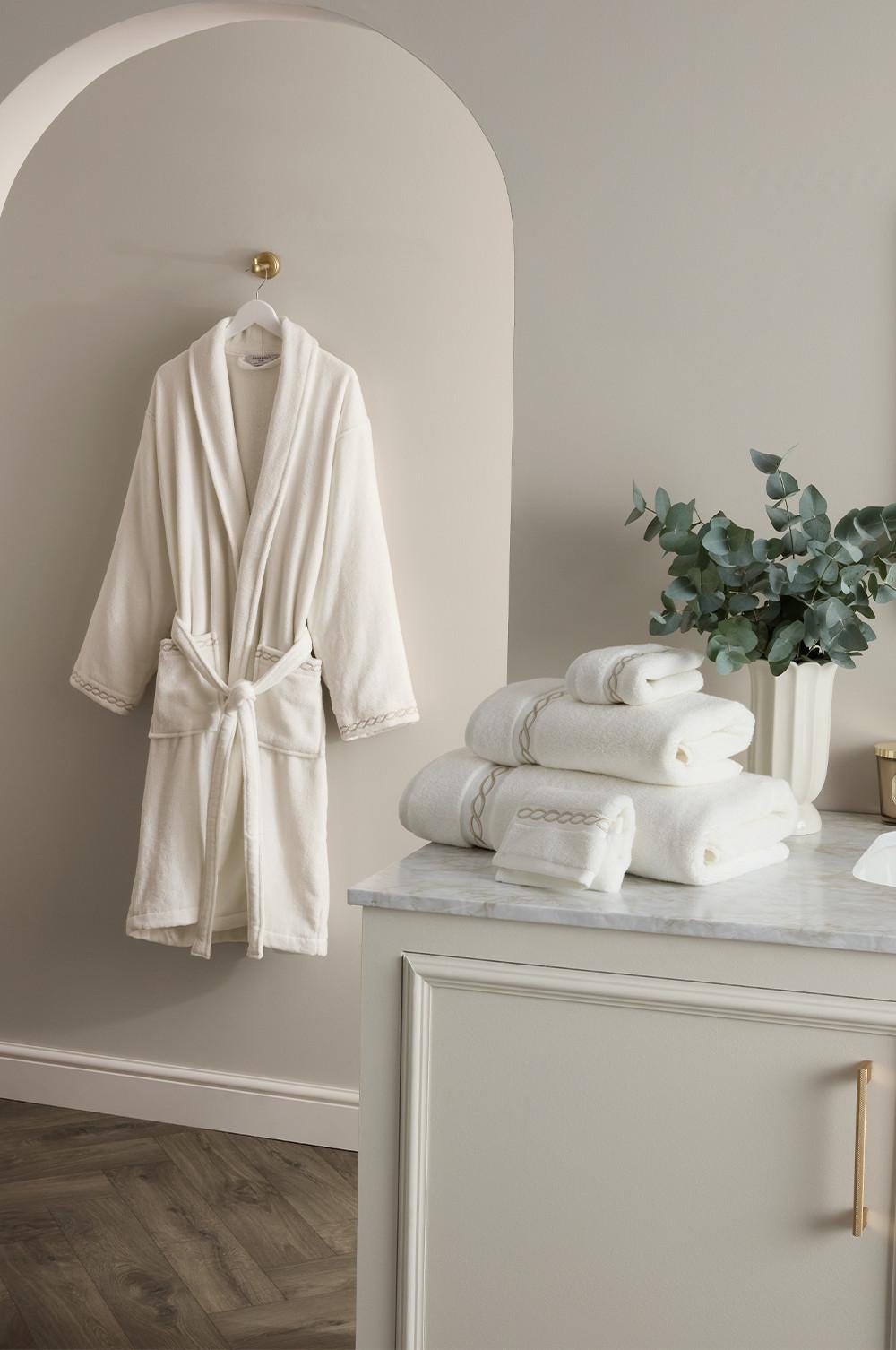 White bathrobe hanging on wall hook with matching towel set and eucalyptus in vase on marble-topped cabinet.