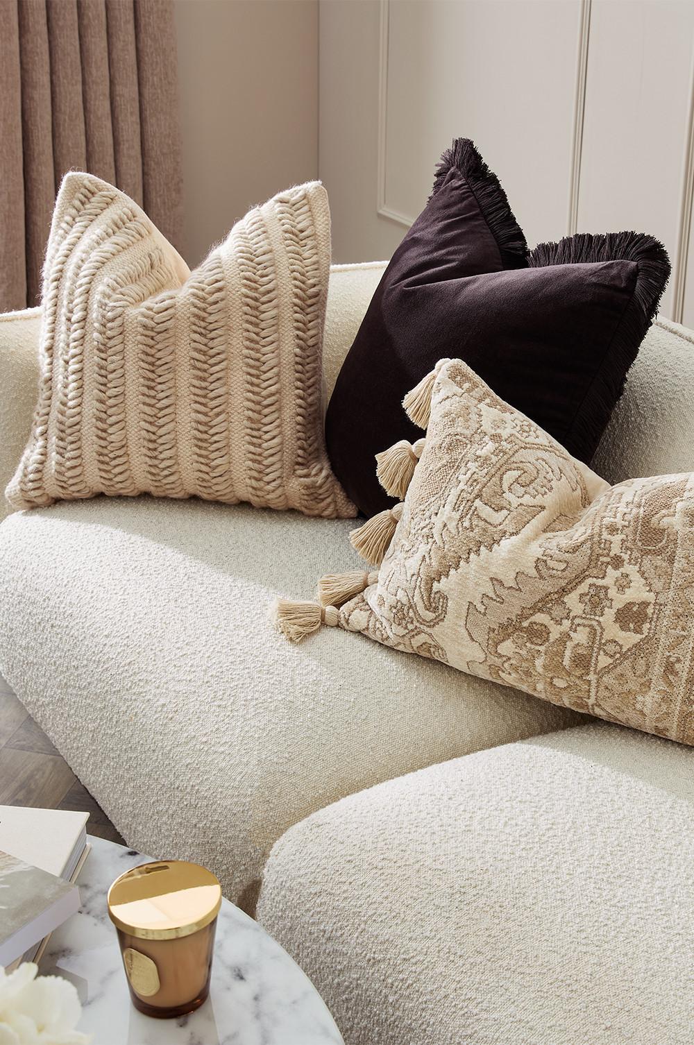 Cream sofa with textured throw pillows in beige, patterned tassel design, and black fringe trim. Elegant home decor arrangement.