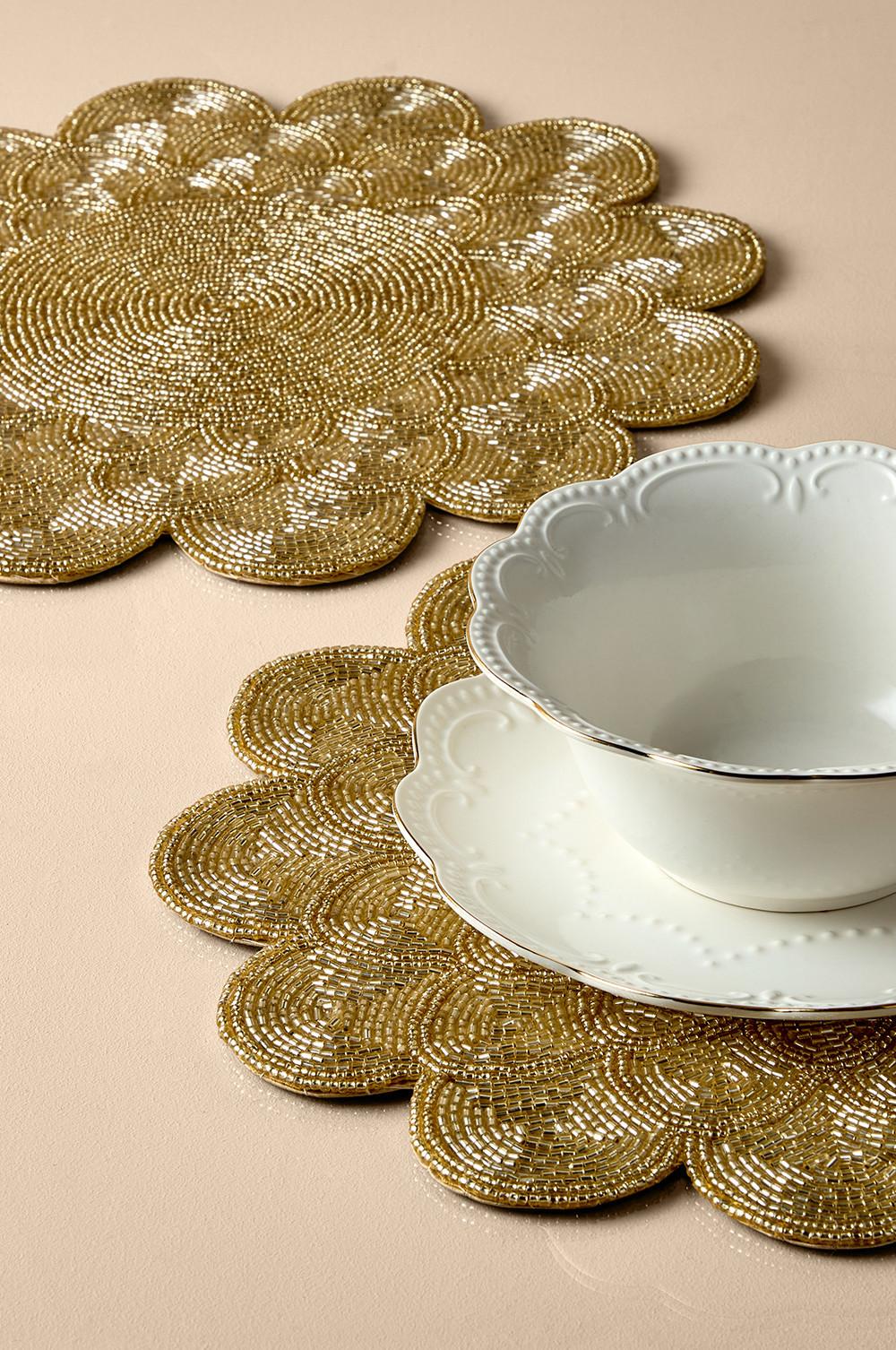 Gold beaded flower-shaped placemats with white porcelain bowl and saucer displayed on cream surface.