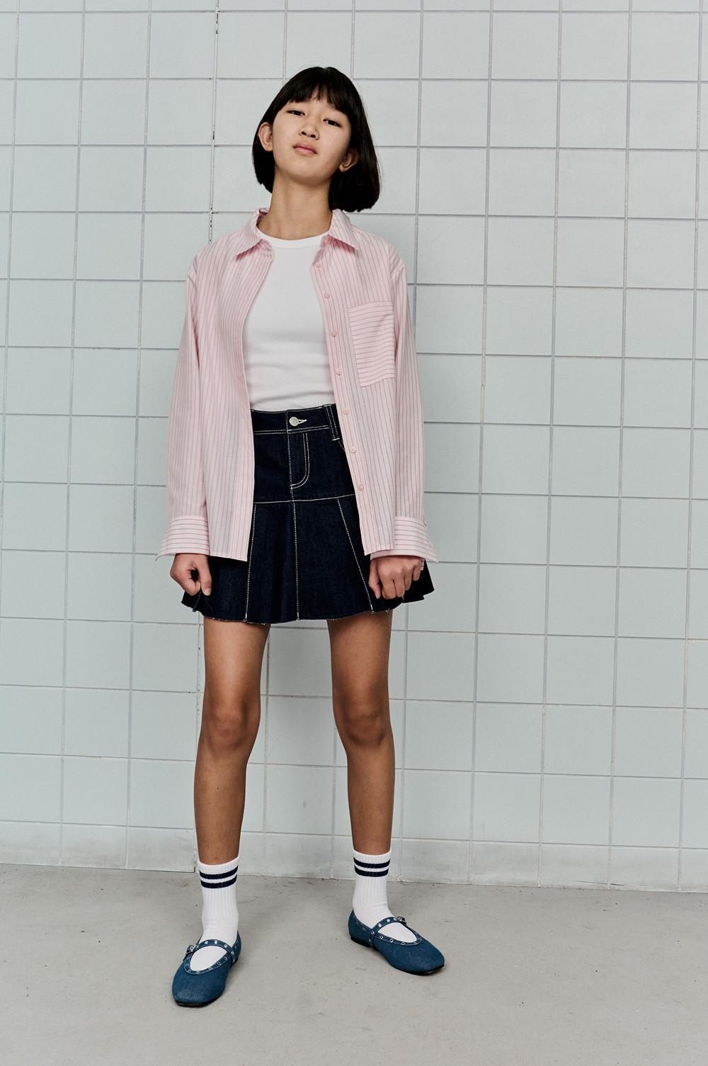 Overshirt a righe rosa con top bianco, minigonna in denim, calzini bianchi a righe e ballerine blu davanti a una parete piastrellata.