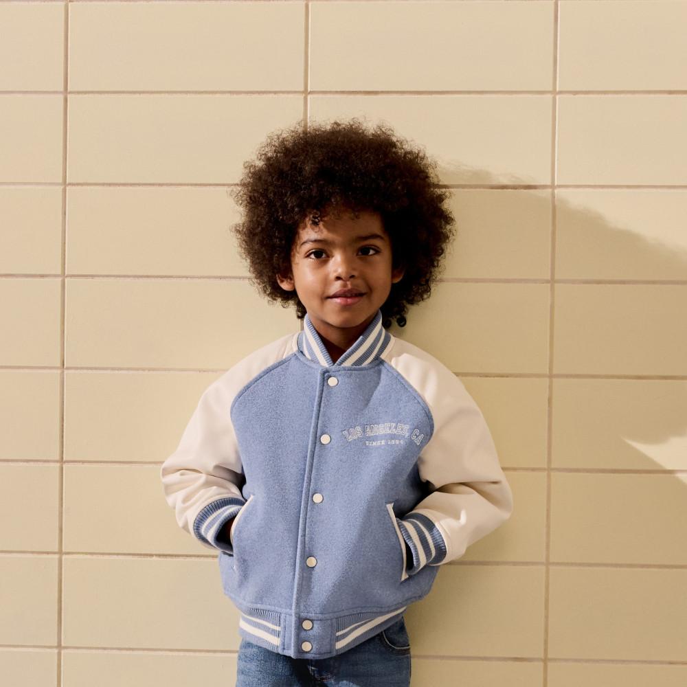 Bambino con una giacca in stile college blu e bianca e capelli ricci davanti a un muro piastrellato.