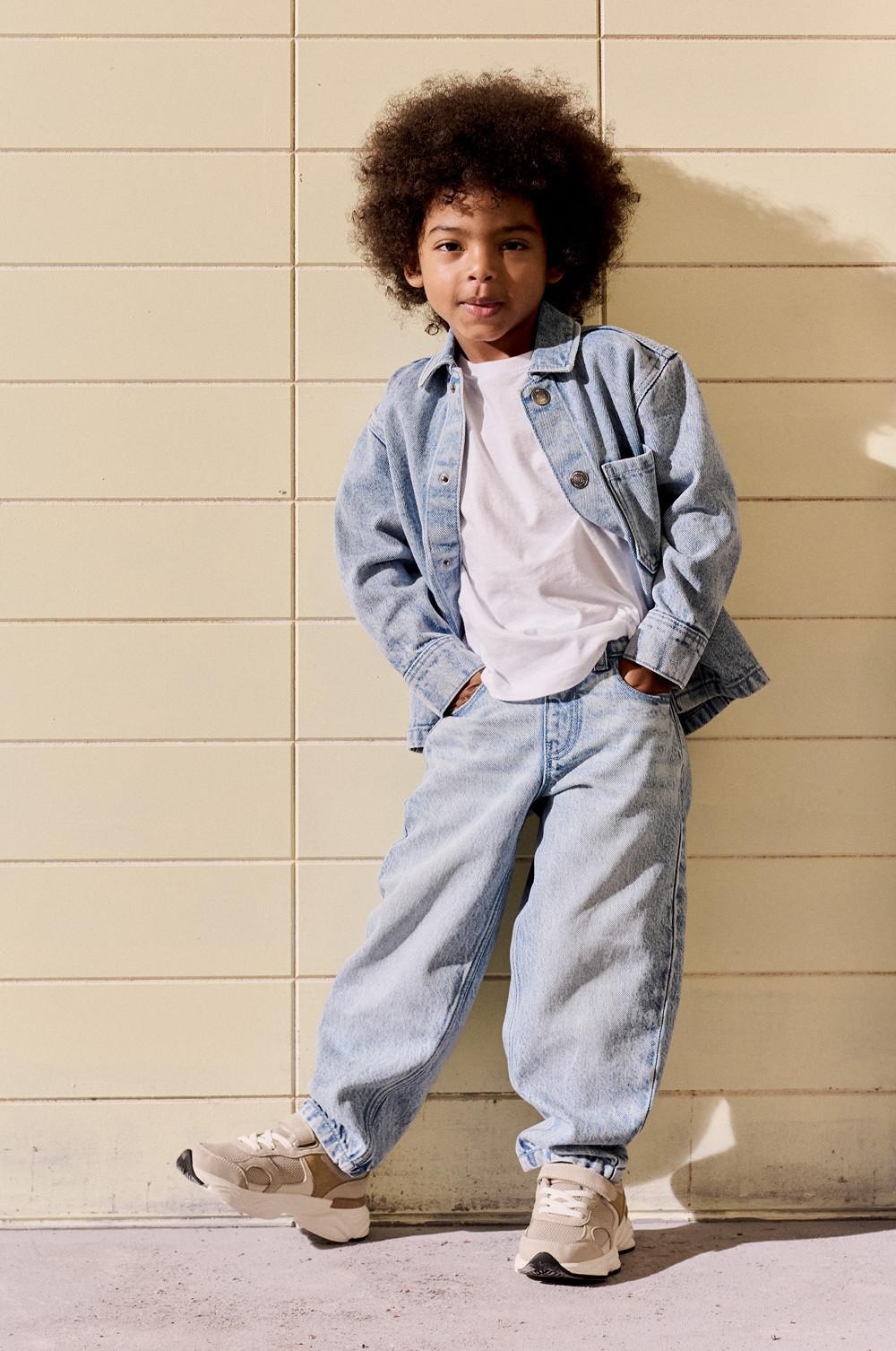 Bambino con giacca di jeans, maglietta bianca, jeans chiari e scarpe da ginnastica beige davanti a una parete piastrellata.