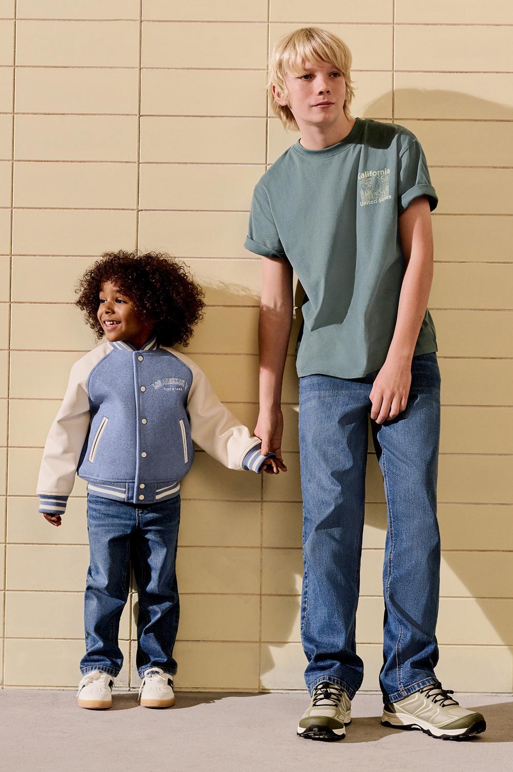 Due bambini vestiti in modo casual: il più piccolo con una giacca blu in stile college, il più grande con una maglietta verde con scritta "California" e un paio di jeans.