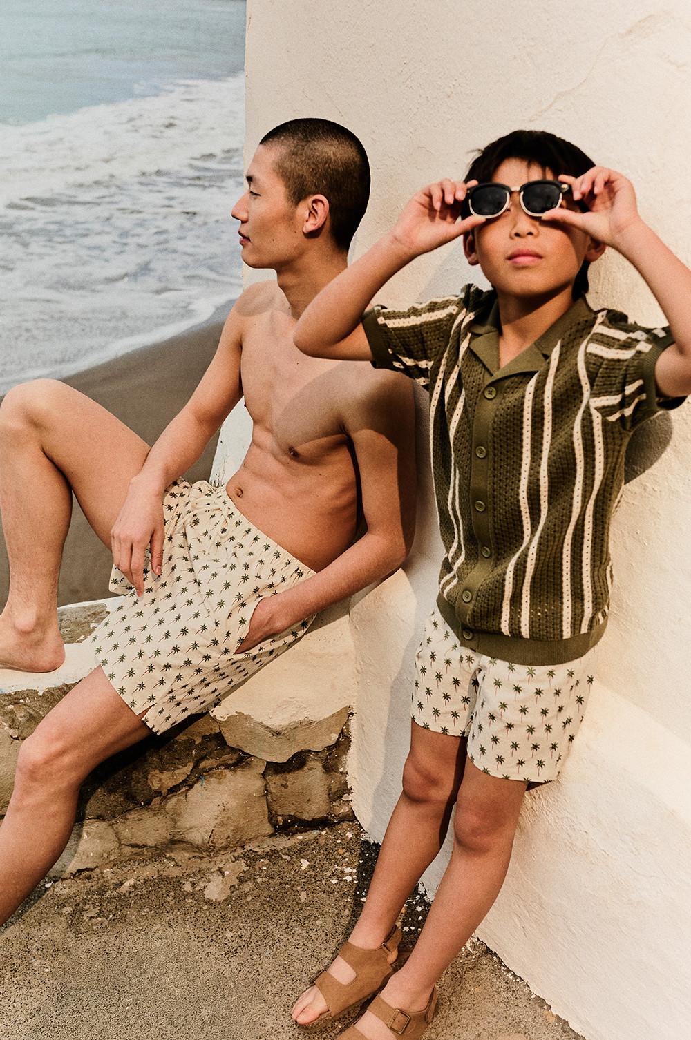 Two men at the beach wearing matching palm tree print shorts and striped shirt with sunglasses.