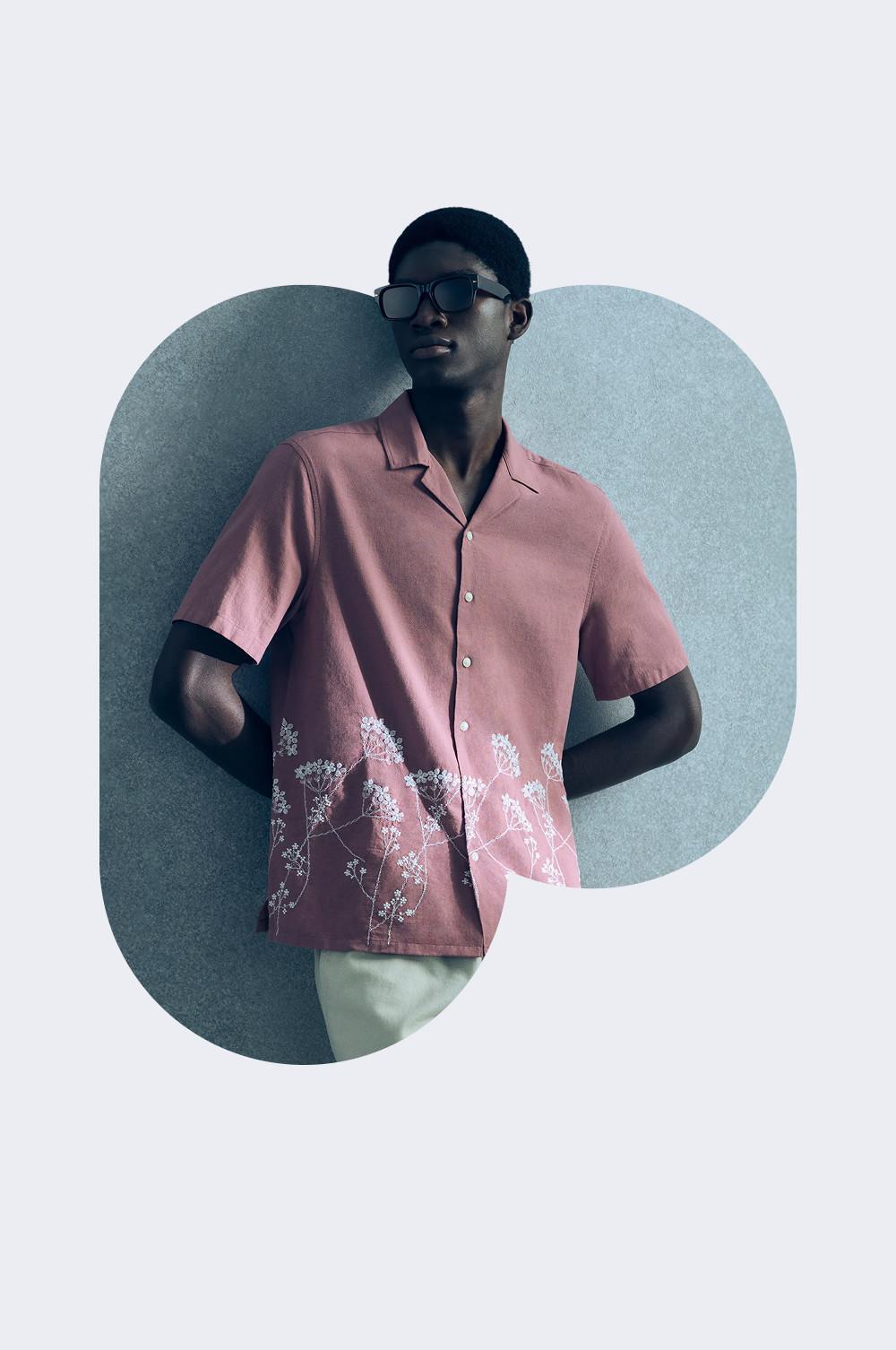 Person mit rosa Kurzarmhemd mit Blumenstickerei und Sonnenbrille vor blauem Hintergrund.
