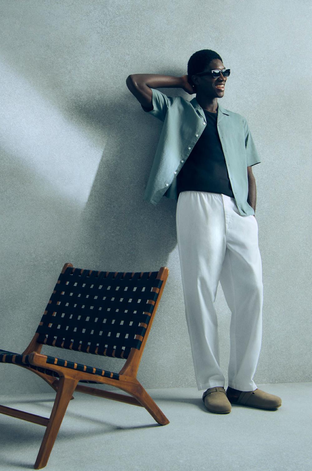 Person mit mintgrünem Hemd über schwarzem Top zu weißer Hose und beigefarbenen Schuhen, neben Holzstuhl stehend.