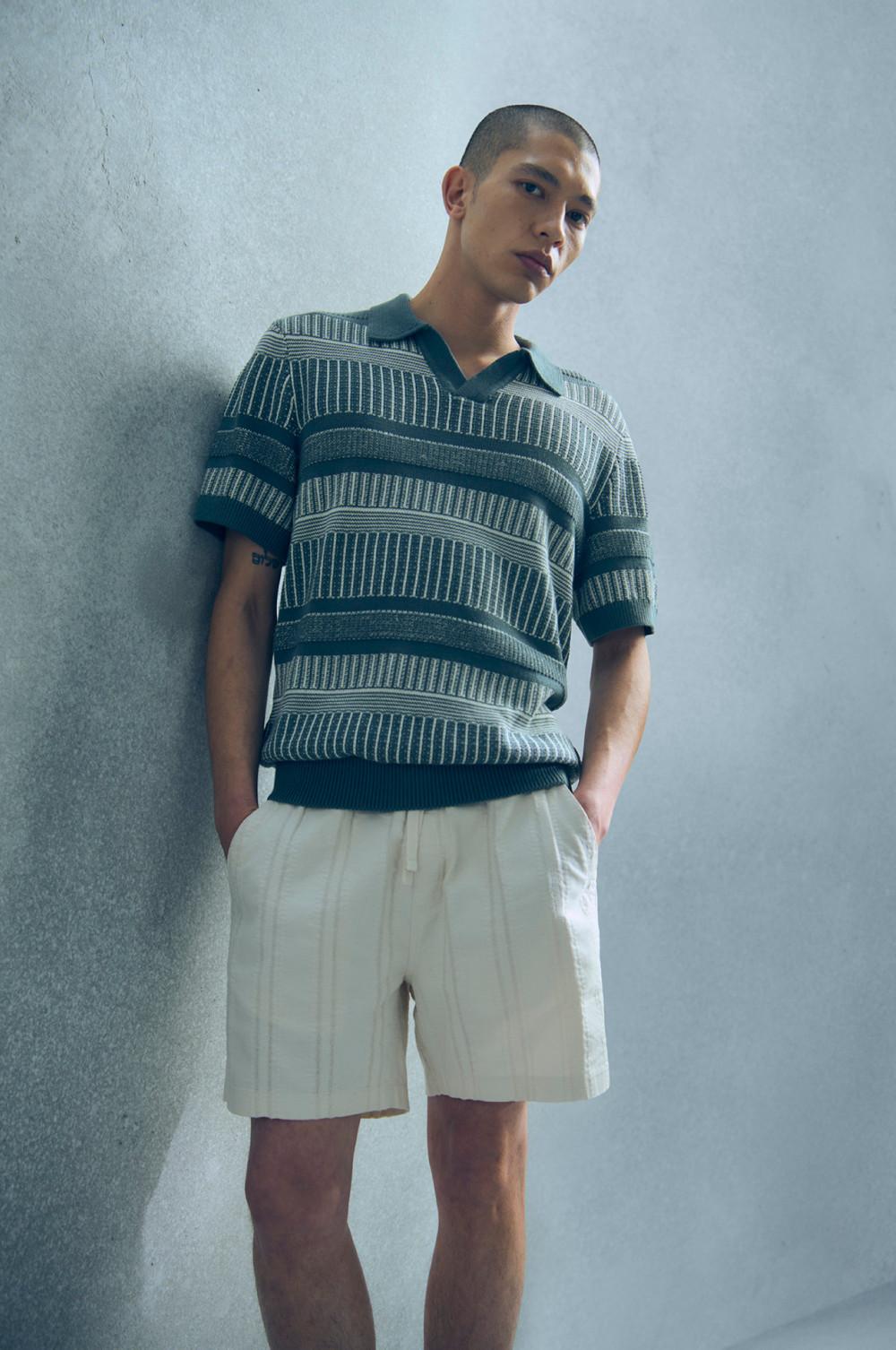 Person mit blaugestreiftem, gestricktem Poloshirt und cremefarbenen Shorts, vor hellgrauer Wand stehend.
