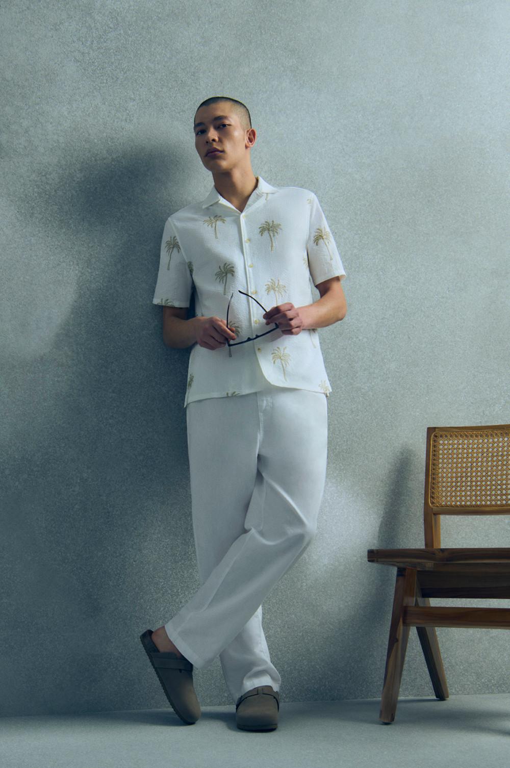 Person mit weißem Kurzarmhemd mit Palmenprint und weißer Hose mit Sonnenbrille in der Hand.