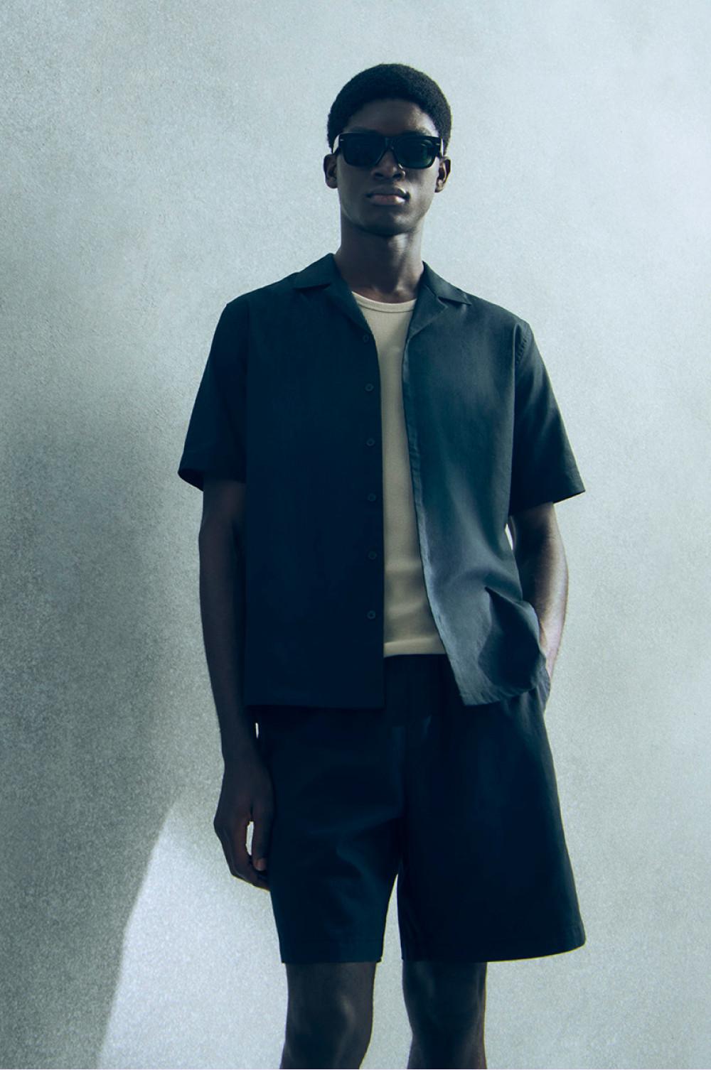 Person mit dunklem Kurzarmhemd über weißem T-Shirt zu passenden Shorts und Sonnenbrille.