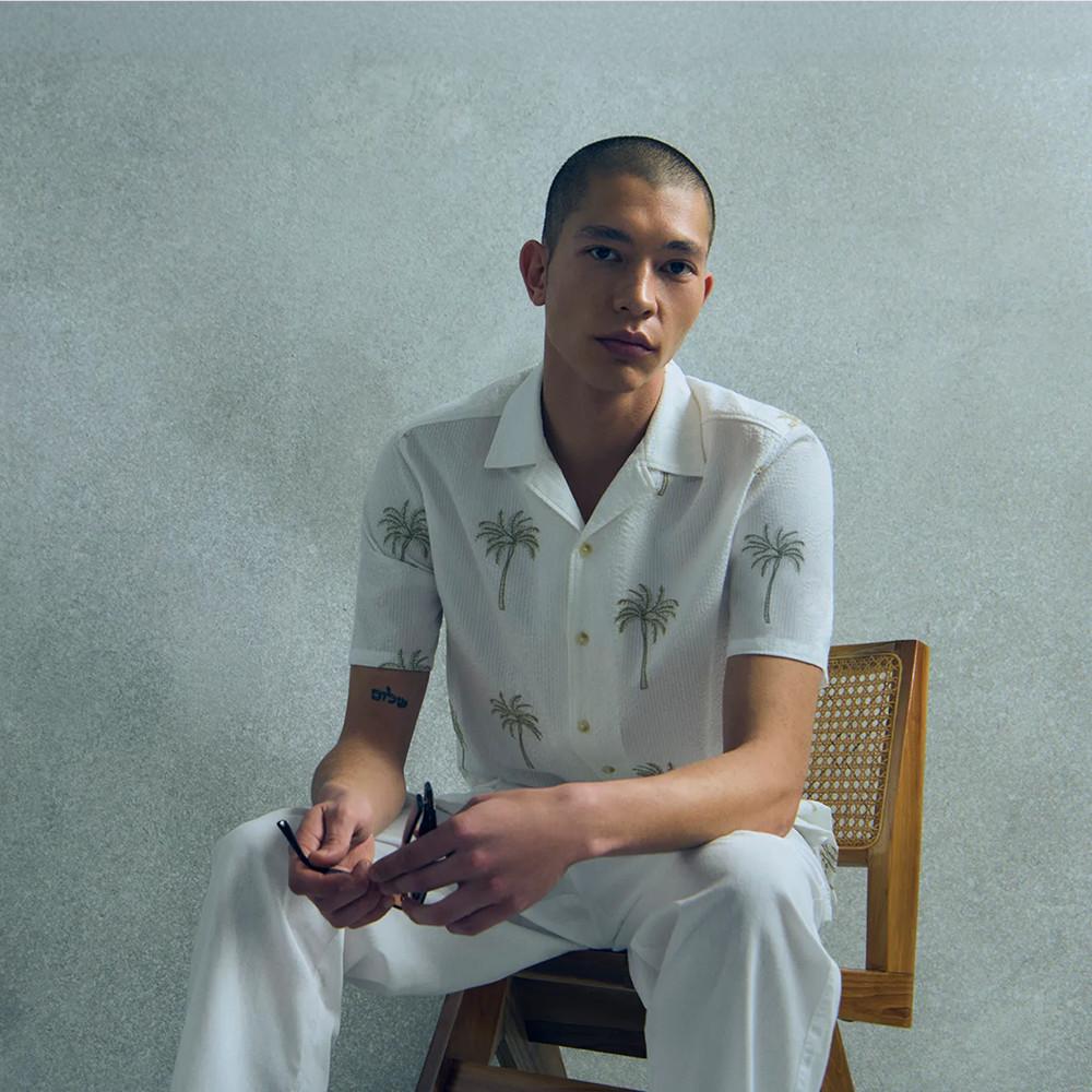 Weißes Kurzarmhemd mit Palmenstickerei und passender Hose, getragen von auf Holzstuhl sitzender Person.