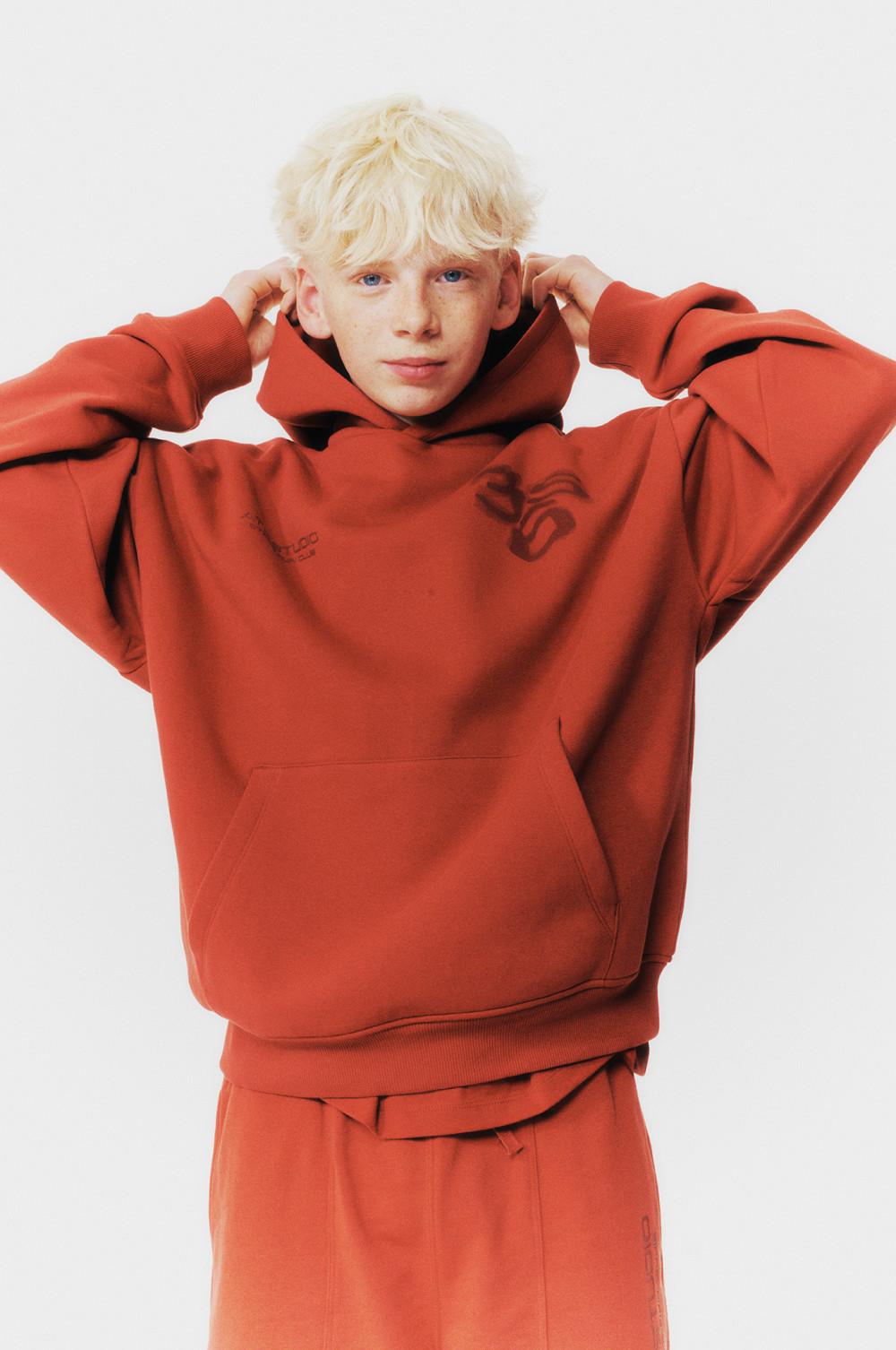 Person wearing an oversized rust-colored hoodie with matching sweatpants, adjusting the hood.