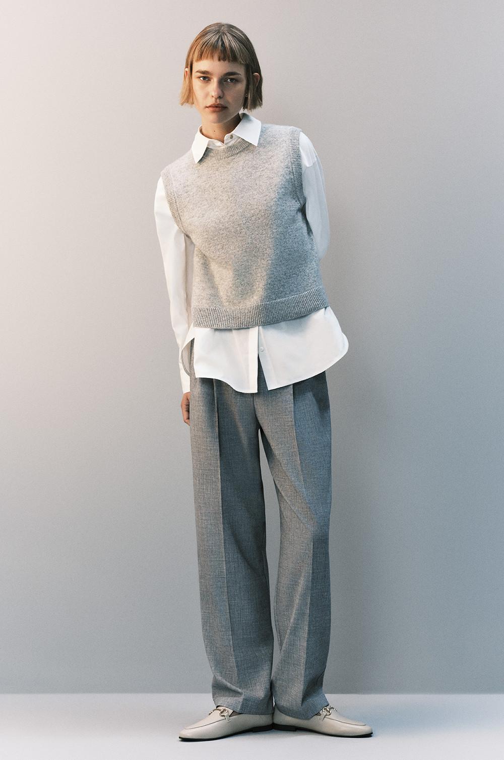 Model wearing grey knit vest over white shirt with wide leg grey trousers and light-coloured loafers.