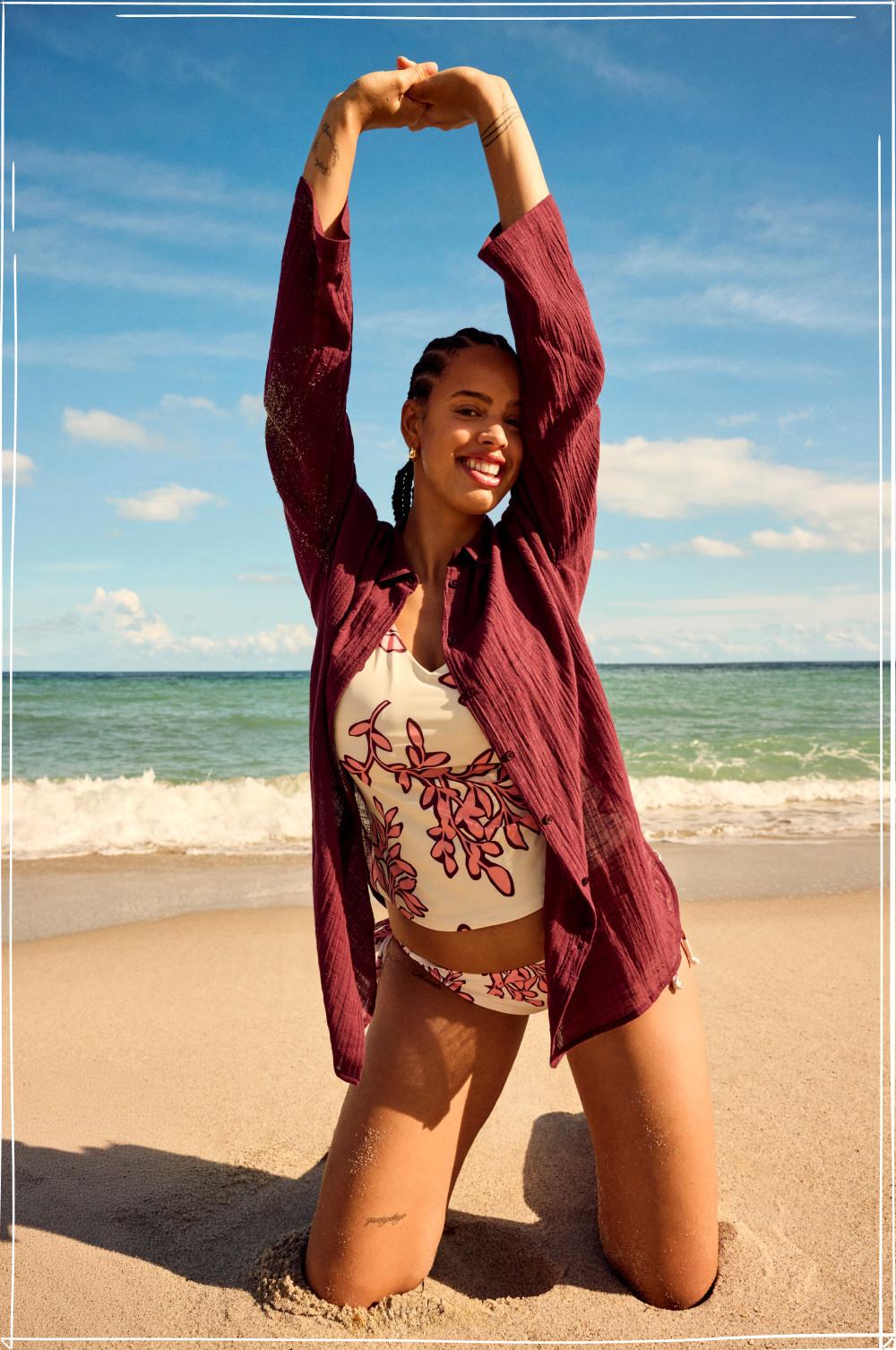 Model kneeling on beach wearing floral swimsuit and burgundy cover-up with arms raised above head.