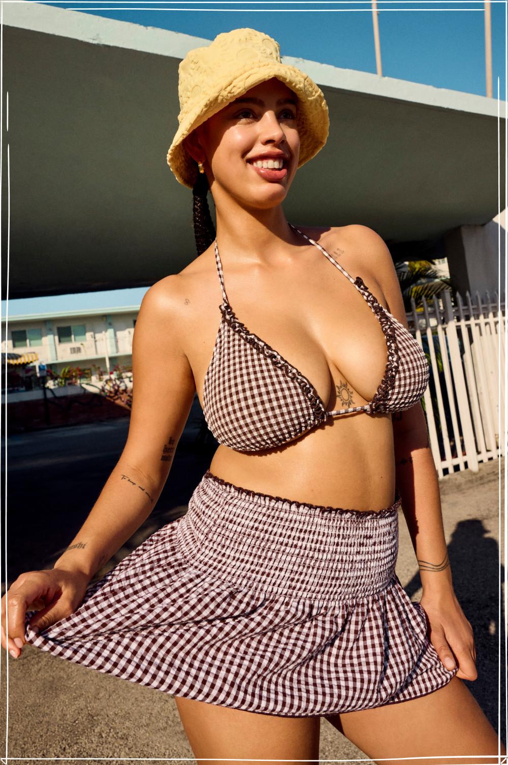 Woman wearing brown gingham bikini top and matching mini skirt with yellow bucket hat outdoors.
