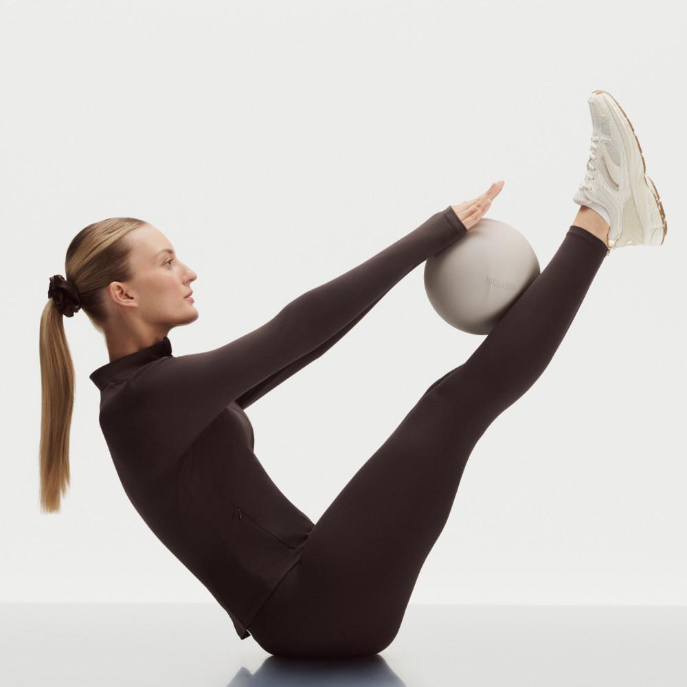 Modella in abbigliamento sportivo nero mentre esegue un esercizio per il core con una palla da ginnastica grigia tra le gambe sollevate e le mani.