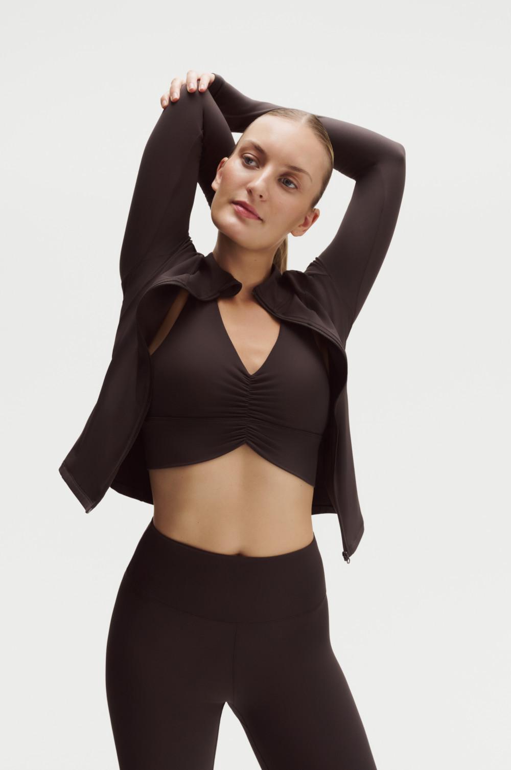 Modella che indossa un completo sportivo nero composto da una giacca con cerniera sopra un top corto arricciato e dei leggings, mentre posa.