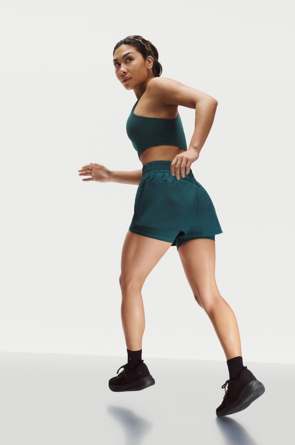 Modella che corre con un reggiseno sportivo verde scuro e pantaloncini abbinati, scarpe da ginnastica nere e una fascia per capelli.
