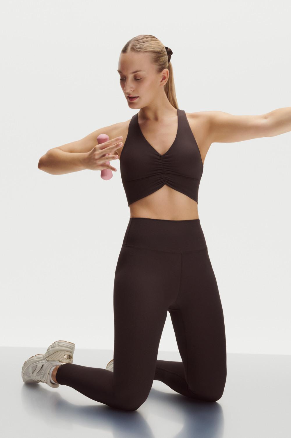 Modella inginocchiata con reggiseno sportivo nero con scollo all'americana e leggings neri, che tiene in mano dei manubri rosa con le braccia in tensione.