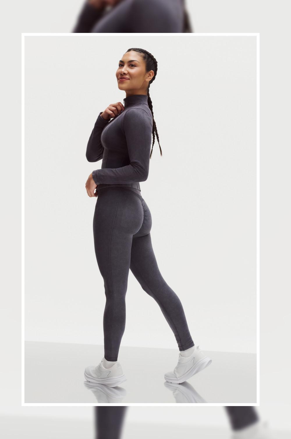 Modella che indossa un completo di abbigliamento sportivo gris marengo composto da maglia a maniche lunghe e leggings, abbinato a scarpe da ginnastica bianche.