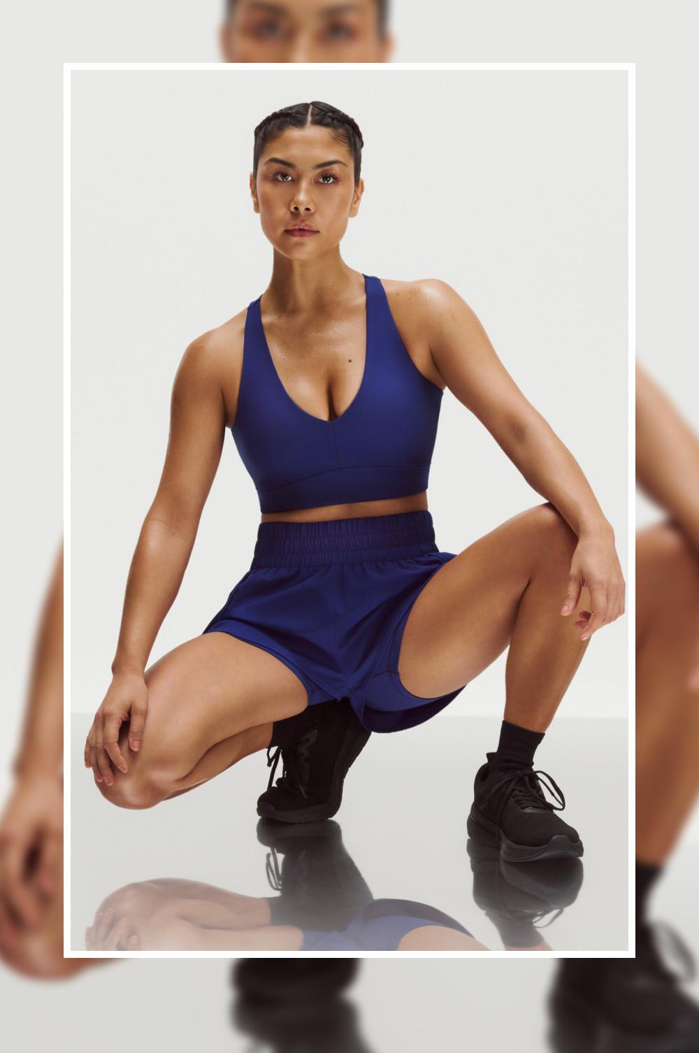 Una modella accovacciata con un reggiseno sportivo blu navy e pantaloncini abbinati, che indossa calzini neri e scarpe da ginnastica nere.