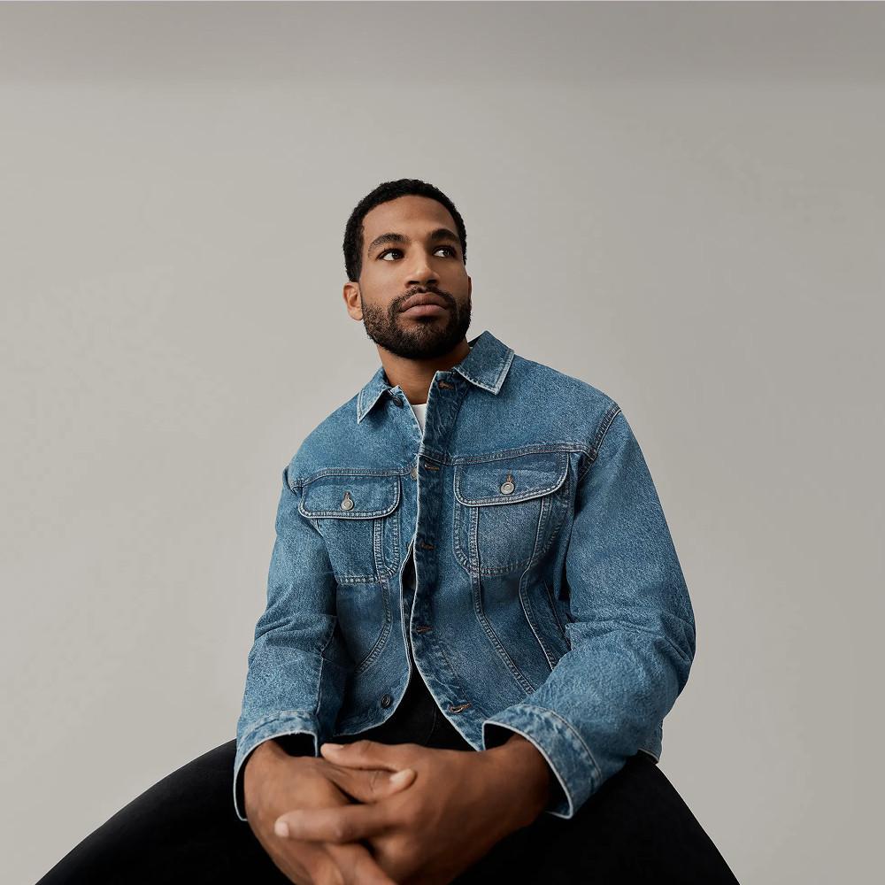 Person wearing denim jacket and jeans with white T-shirt, seated on fabric rolls, wearing black loafers.