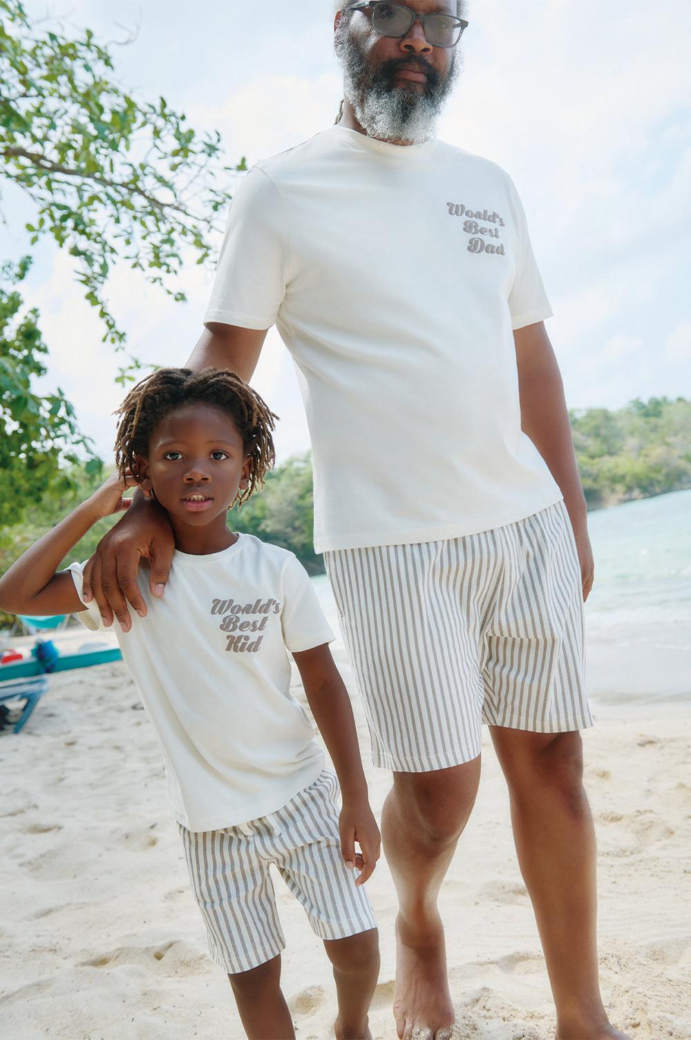 Matching white T-shirts with "World's Best Dad" and "World's Best Kid" text, paired with striped shorts on a beach setting.