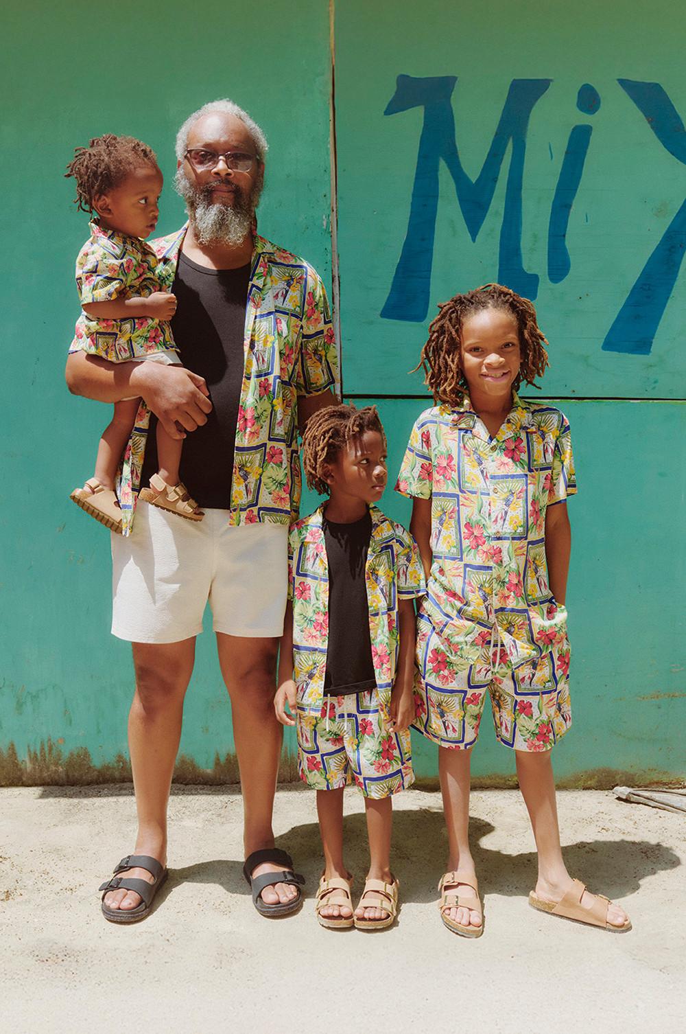 Family wearing matching colorful tropical print shirts and shorts standing against a turquoise wall with blue text.