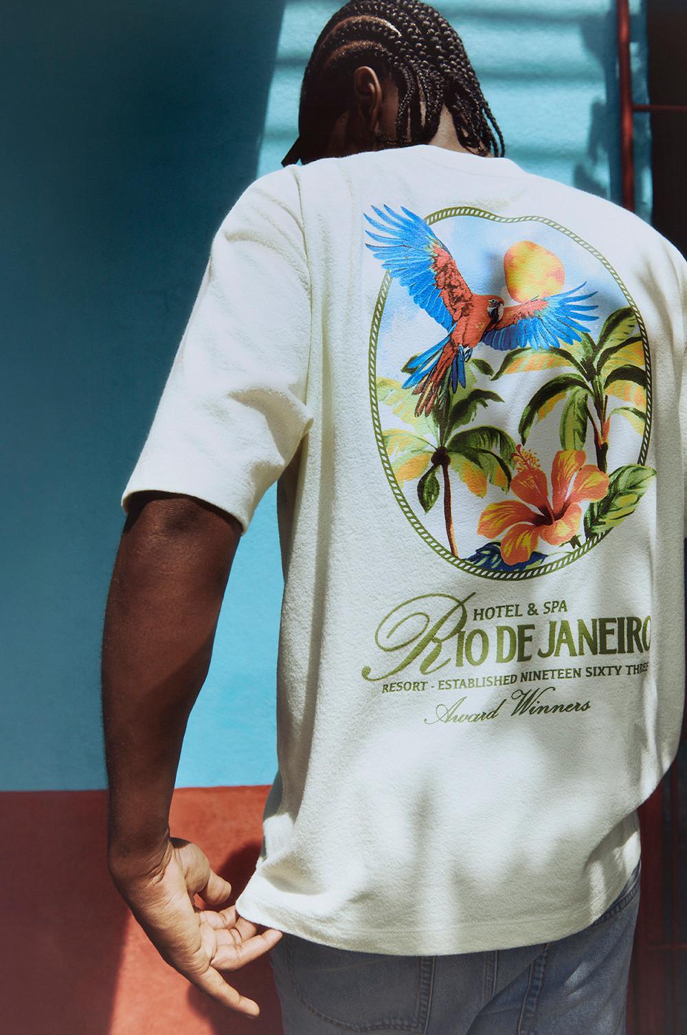 White T-shirt featuring tropical design with parrot, palm trees and flowers, branded with Rio De Janeiro Hotel & Spa logo on the back.