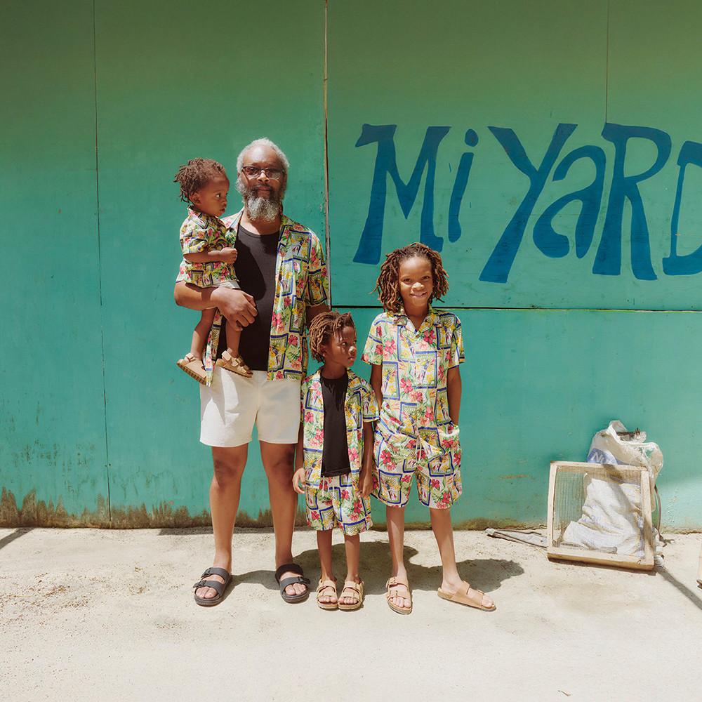 Family wearing matching colourful tropical print shirts and shorts standing against a turquoise wall with blue text.