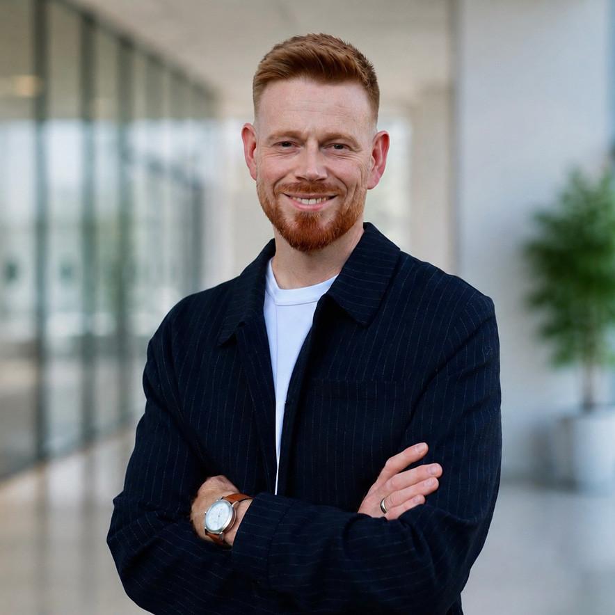 A smiling man with short ginger hair and a beard, wearing a t-shirt and a dark pinstriped jacket.