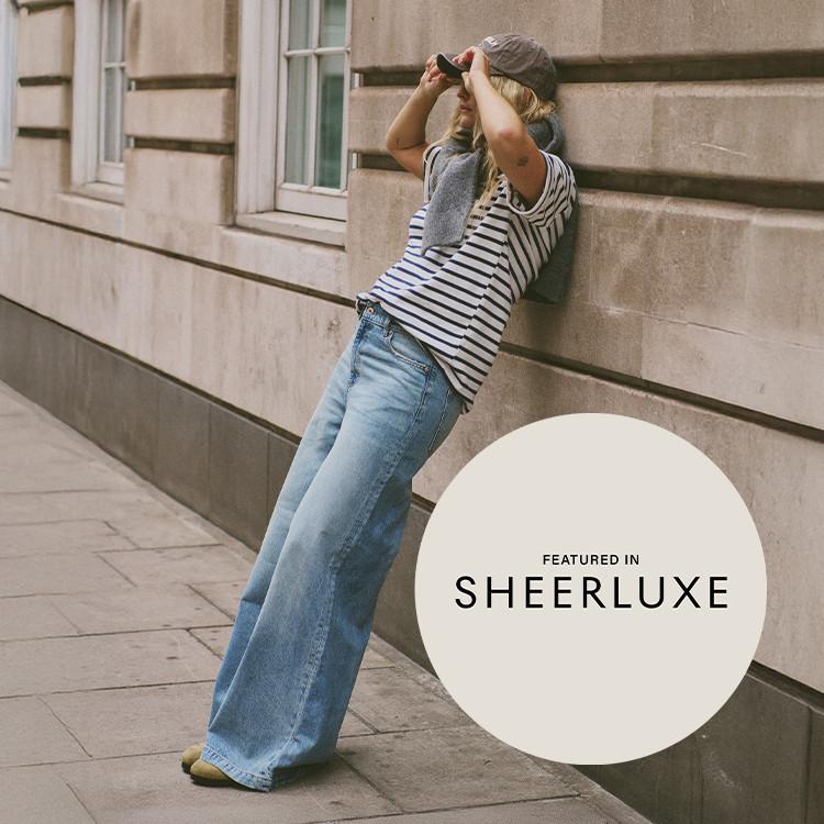 Person wearing striped top and wide-leg jeans leaning against building wall, featured in SHEERLUXE.