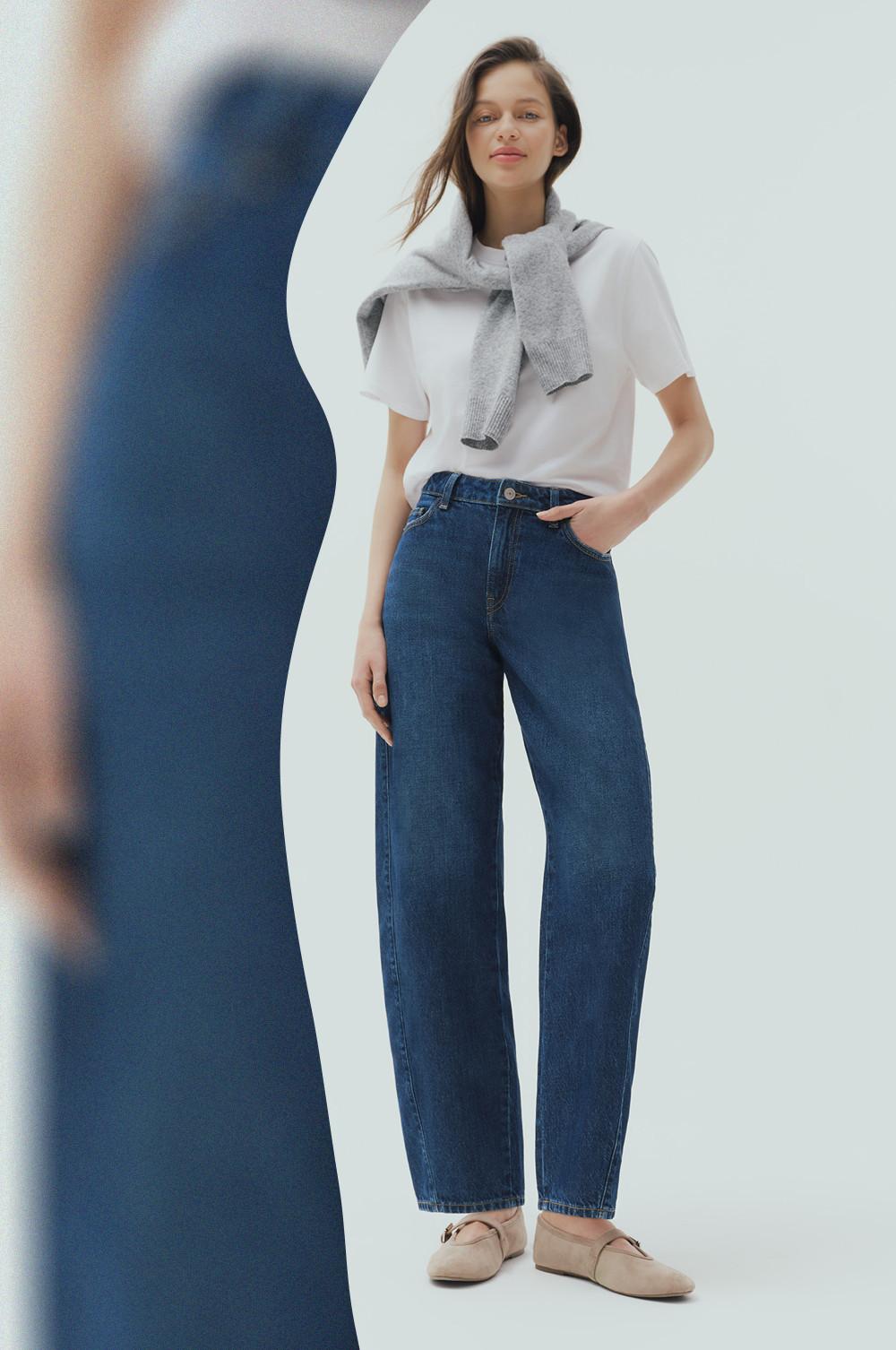 Model wearing white T-shirt with gray sweater draped over shoulders, blue wide-leg jeans and beige loafers.