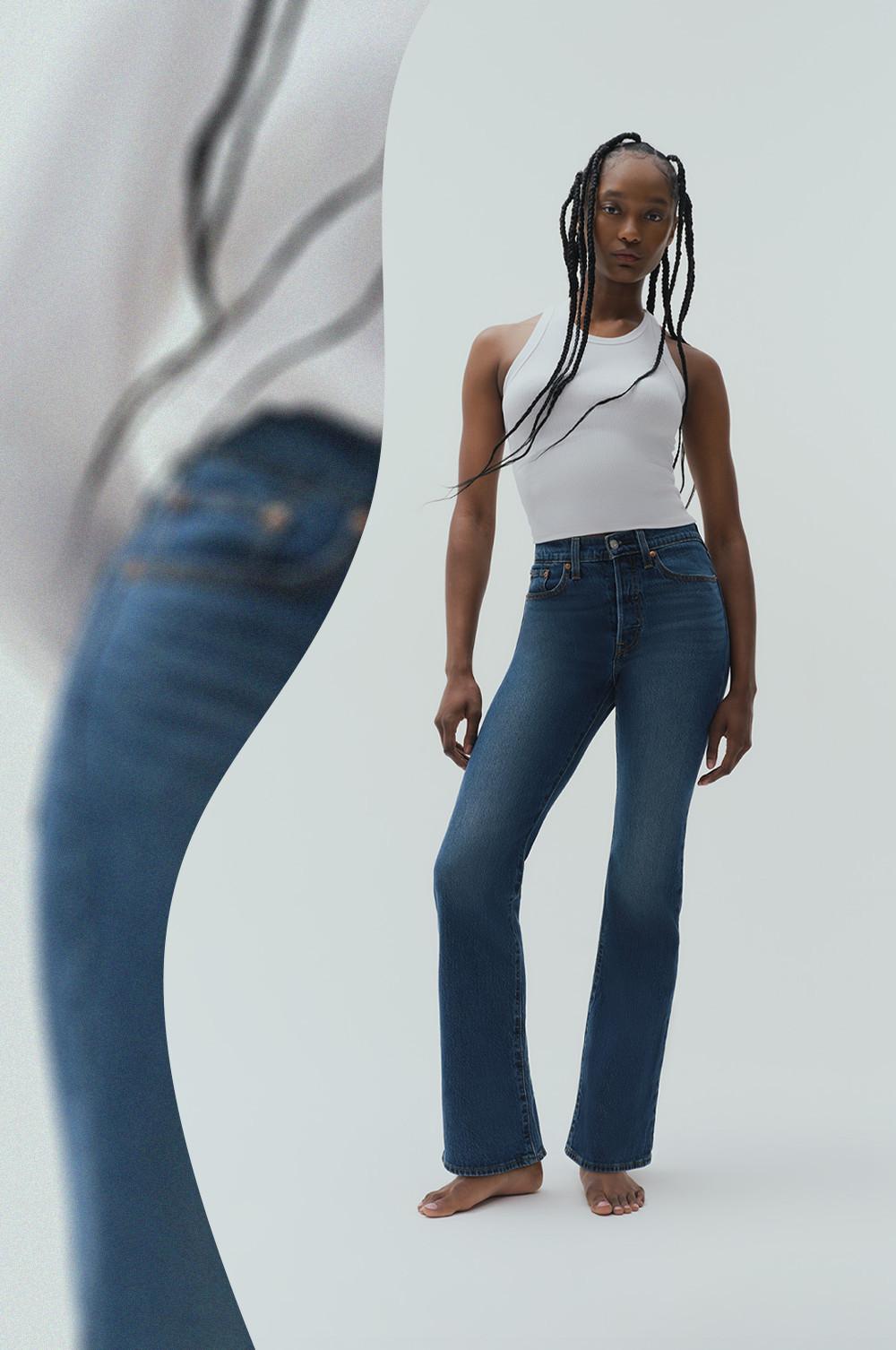 Model wearing white sleeveless top and blue bootcut jeans standing barefoot.