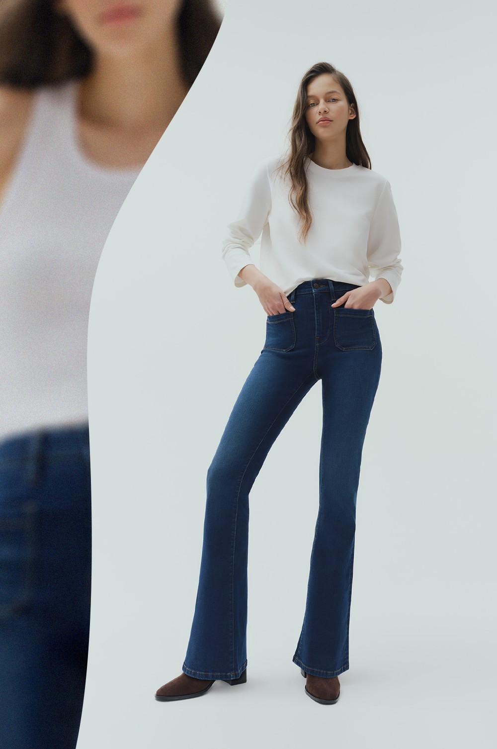 Model wearing a white long-sleeved top with dark blue flared jeans and brown shoes, hands in pockets.