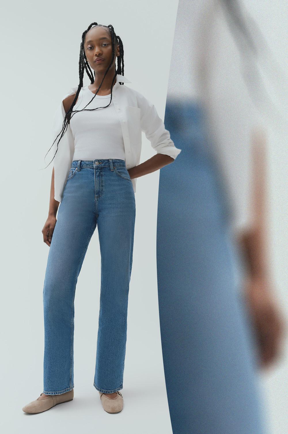 Model wearing white off-shoulder top, cream blazer and blue straight-leg jeans with beige ballet flats.
