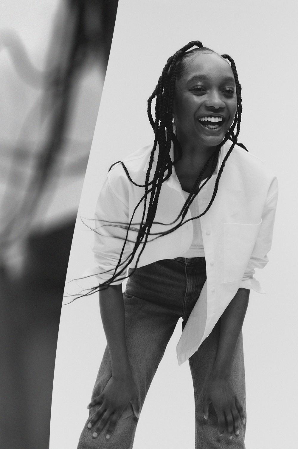 Model smiling wearing a white long-sleeved top with dark jeans and long braided hair.