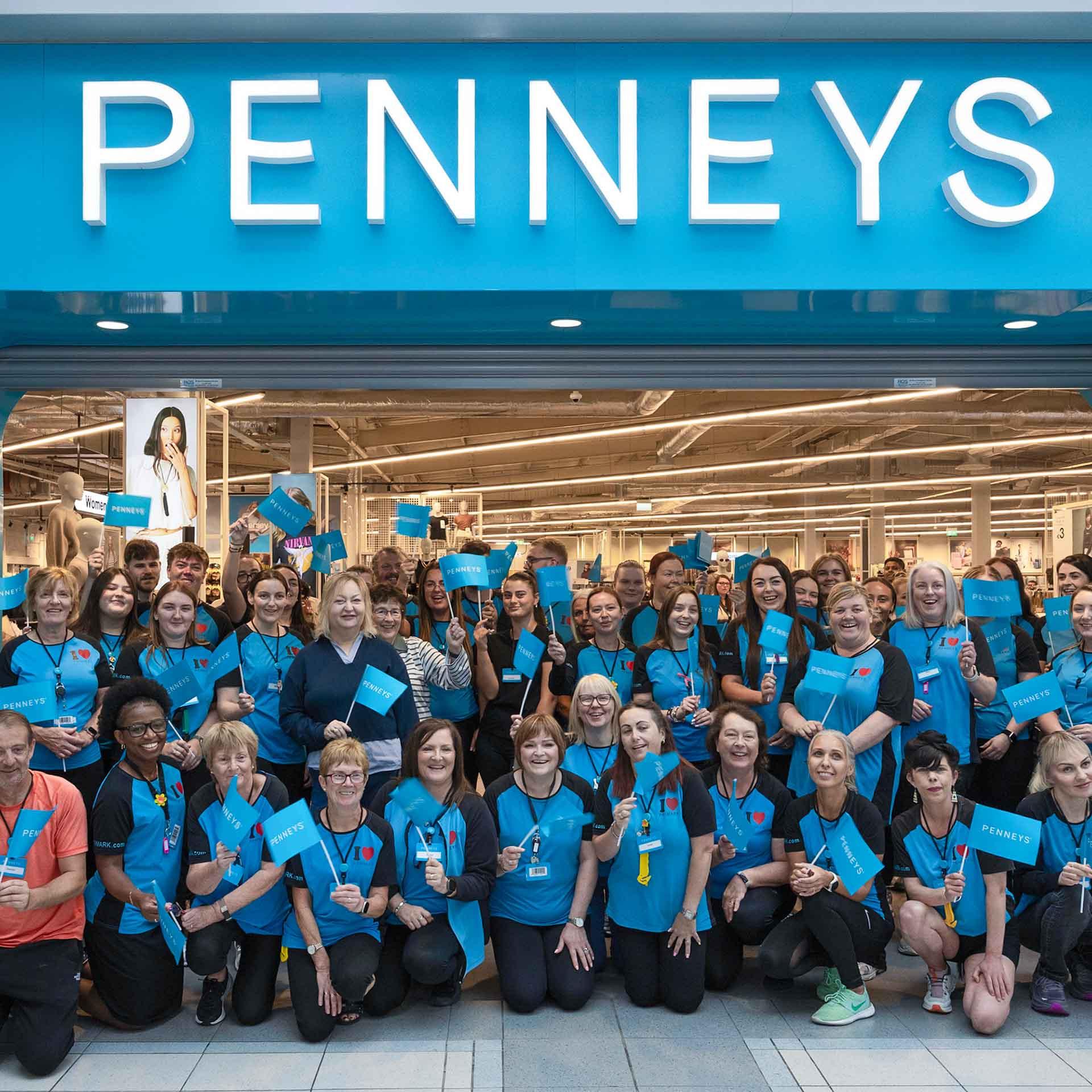 A large group of Penneys employees posing in front of the store entrance, holding Penneys flags.