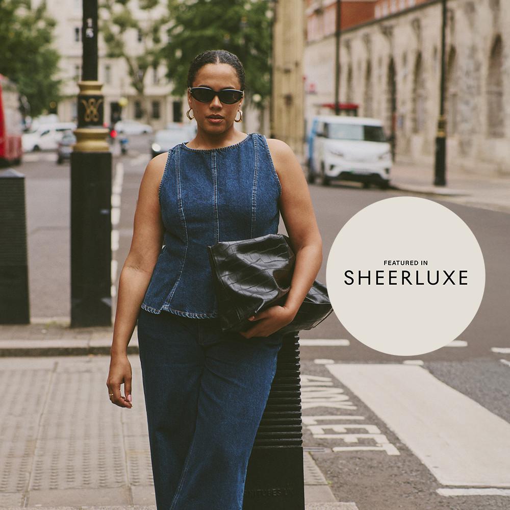 Person wearing stylish denim outfit with sunglasses, carrying black clutch bag on city street. Featured in SHEERLUXE.