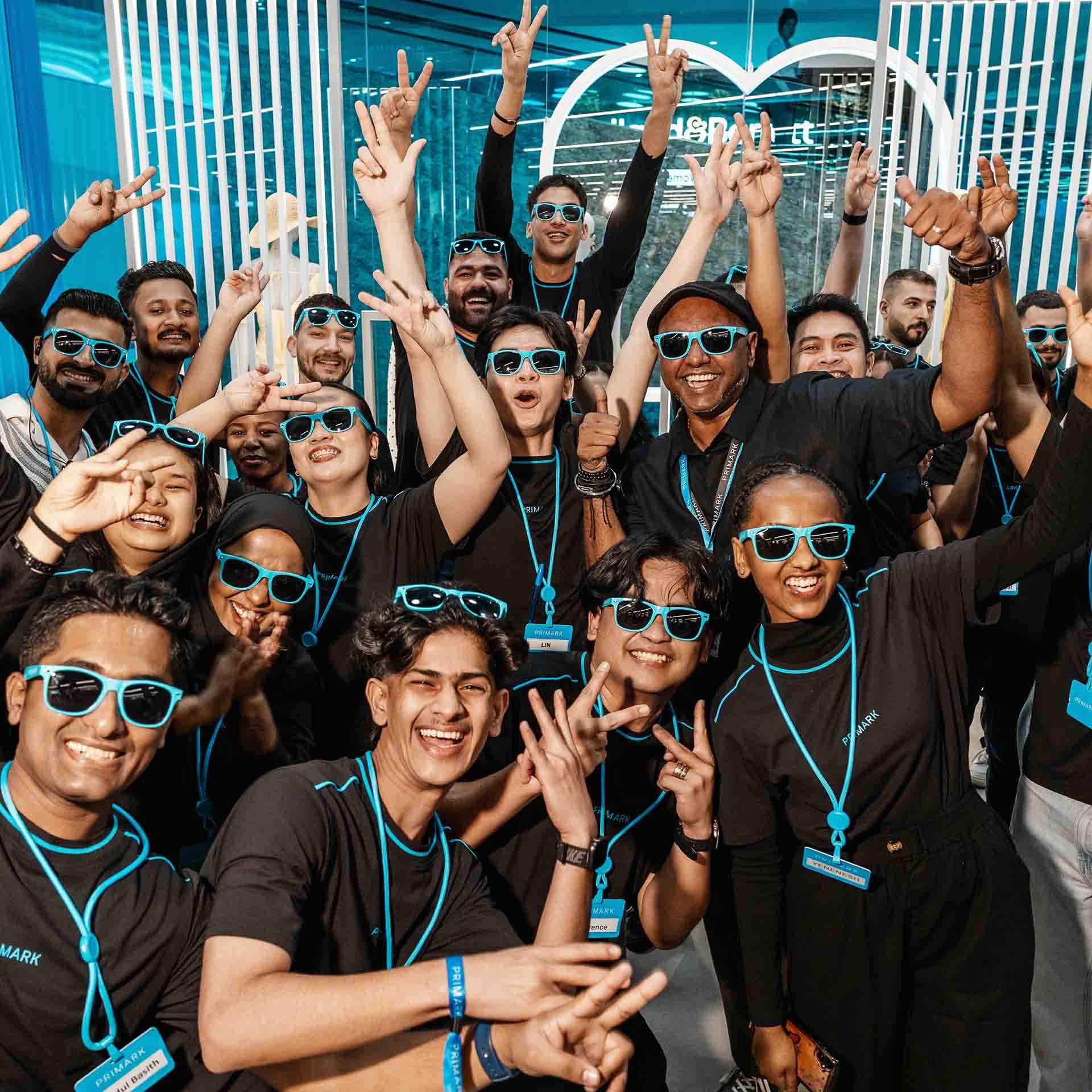 A group of people wearing blue sunglasses & black shirts joyfully pose with peace signs & smiles.