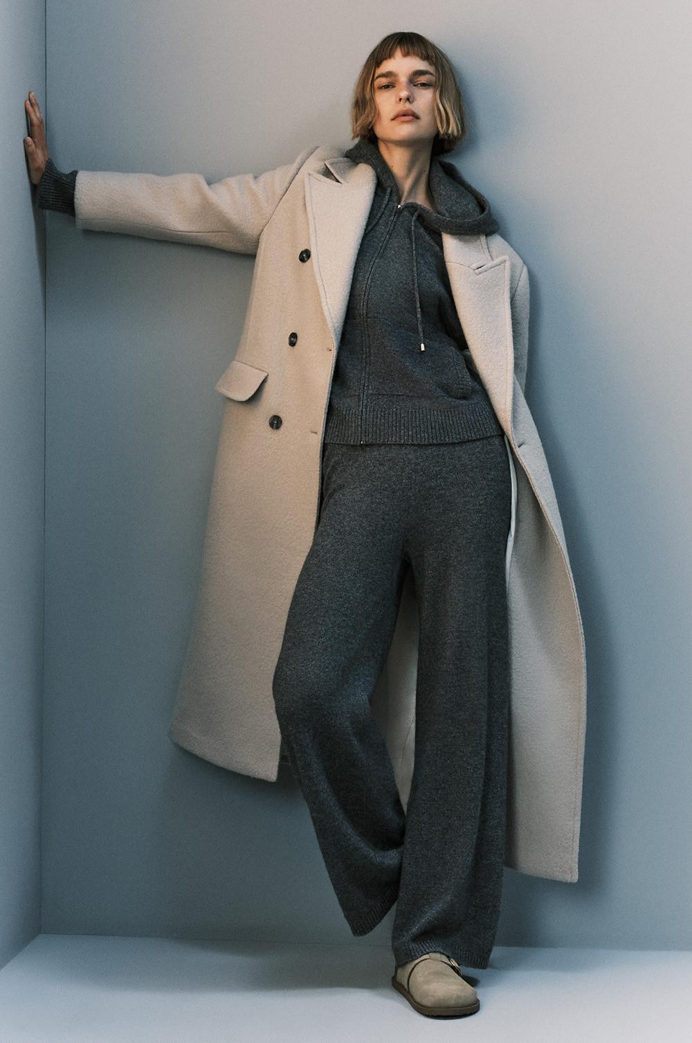 Person wearing cream wool coat over grey hoodie and matching wide-leg trousers with beige shoes.
