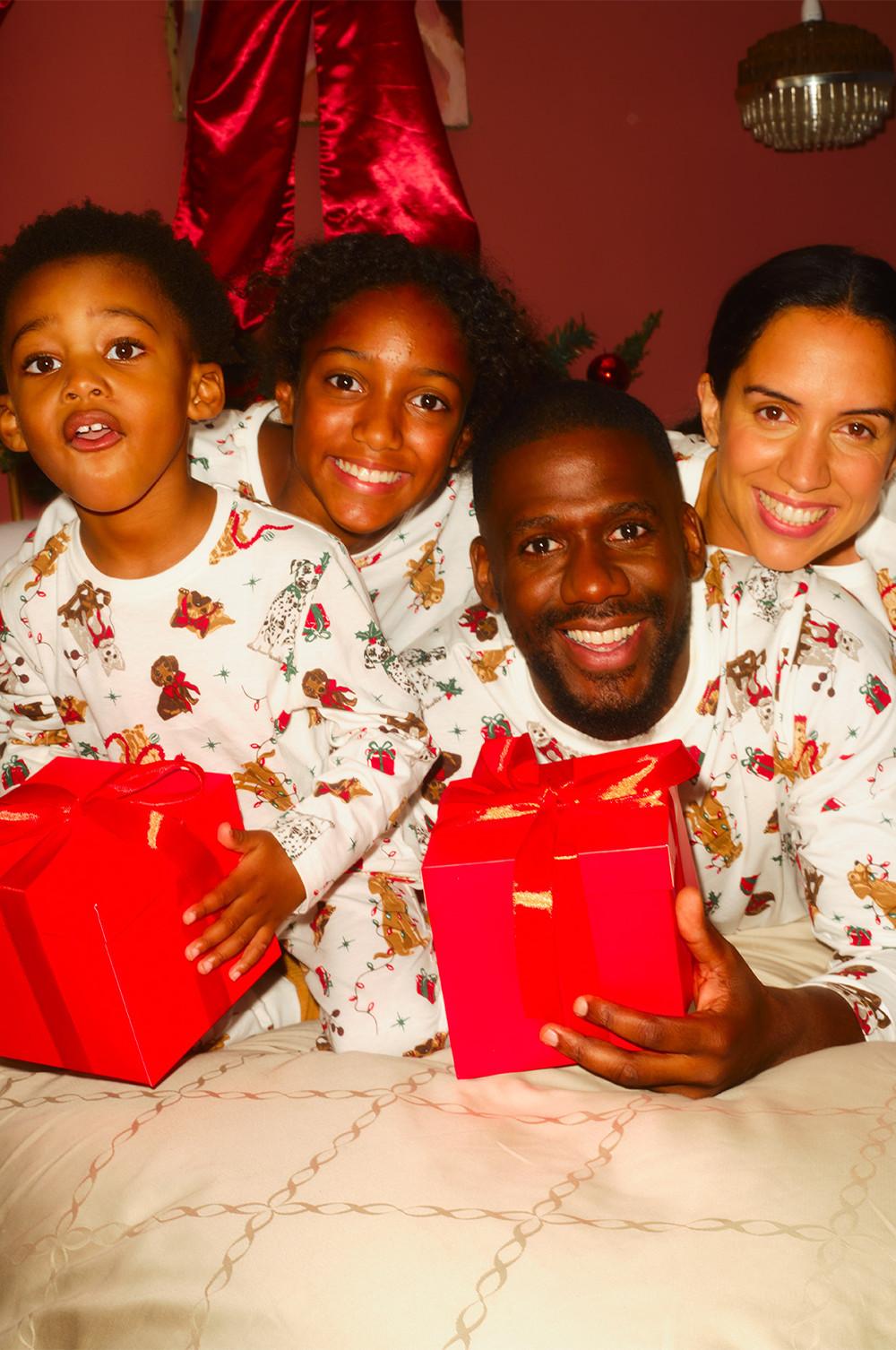 Family wearing matching Christmas pyjamas with gingerbread pattern, holding red gift boxes.