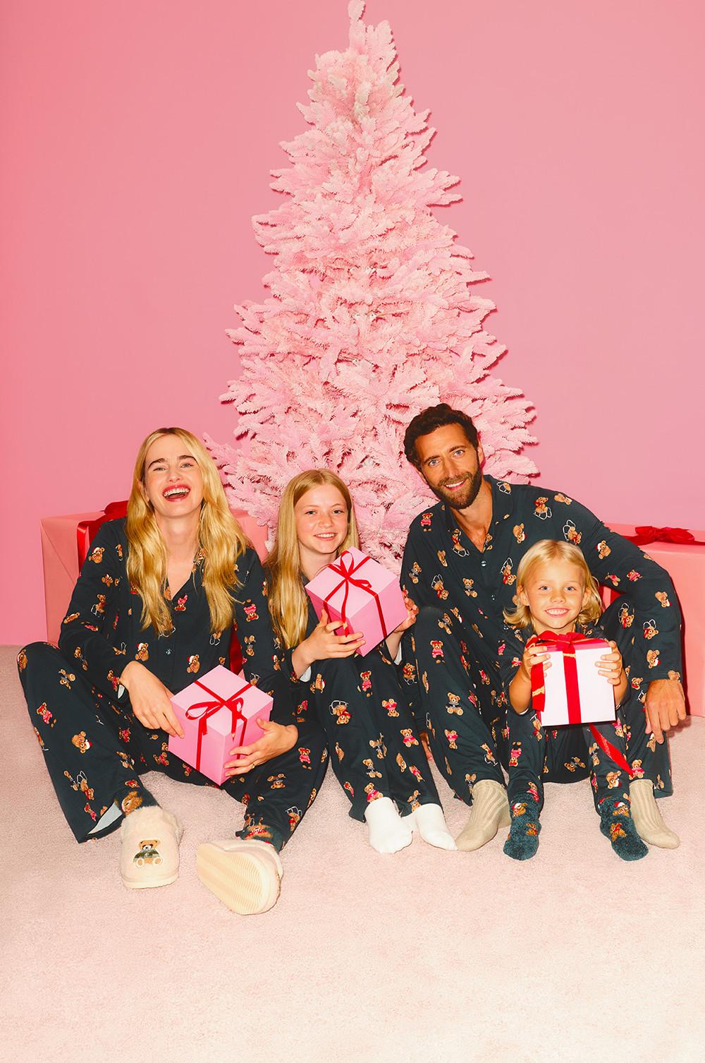 Familia vestida con pijamas a juego con estampado de ositos de peluche posa sosteniendo cajas de regalo delante de un árbol de Navidad rosa.