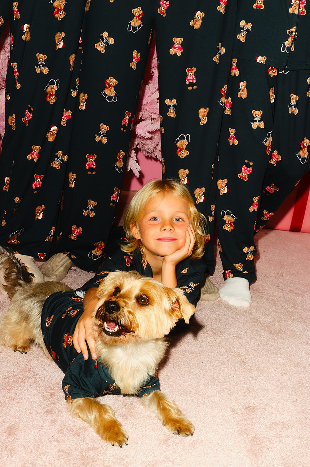 Small dog wearing teddy bear patterned pyjamas next to child in matching outfit under festive curtains.